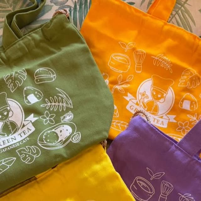 Four Utsuwa Green Tea original tote bags in olive, orange, purple and yellow, printed with the café's mascot logo