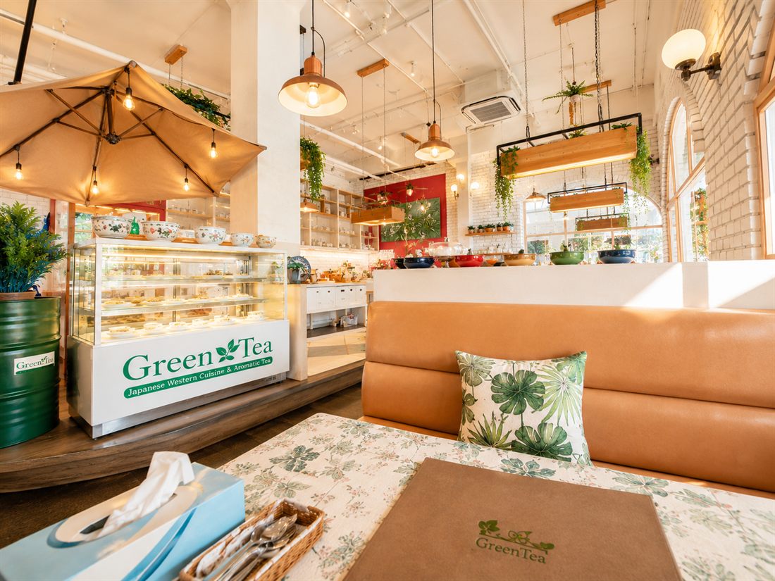 The Green Tea cake display case beside a tan leather booth with a tropical-leaf cushion
